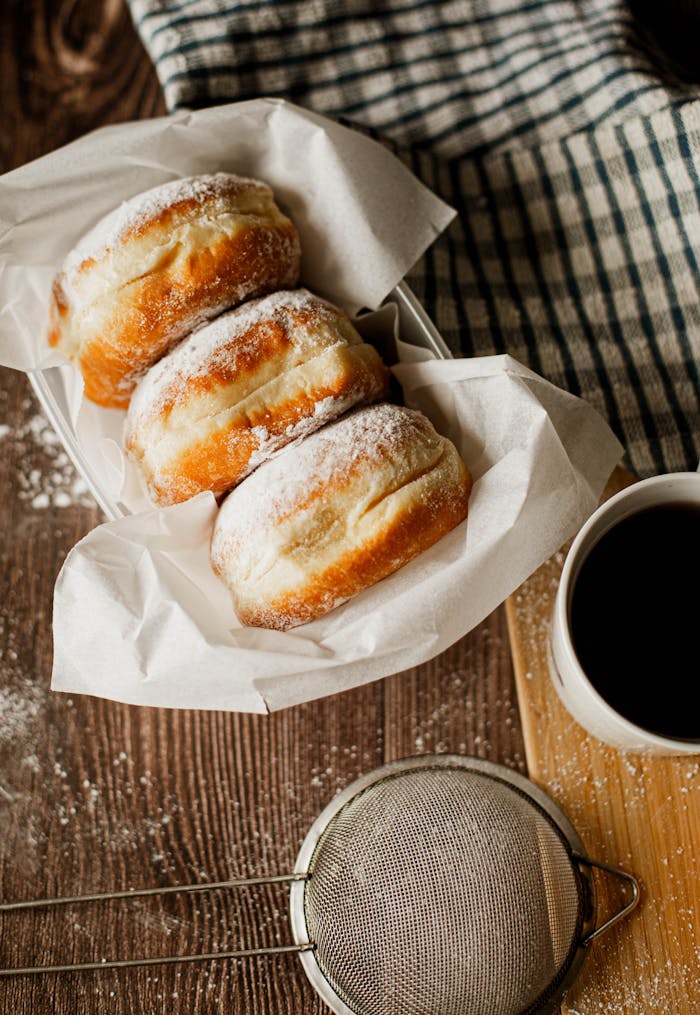 Three donuts with powdered sugar paired with a cup of black coffee, captured in warm natural light.